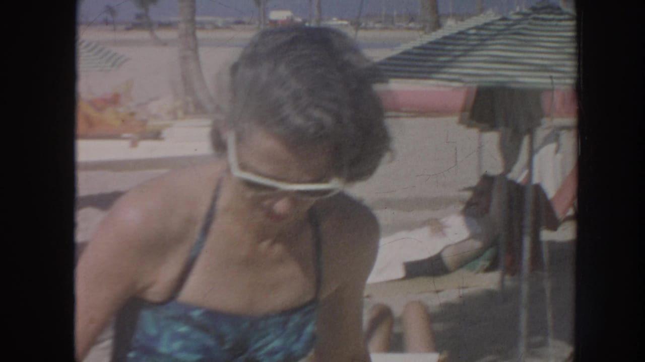 1961 Hawaii home-movie frame of a woman in white sunglasses and a blue patterned swimsuit sitting on a beach chair beside a striped umbrella.