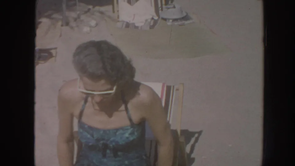 1961 Hawaii beach frame with a woman in a blue patterned swimsuit seated in a chair, seen from above with sand, chairs, and beachgoers nearby.