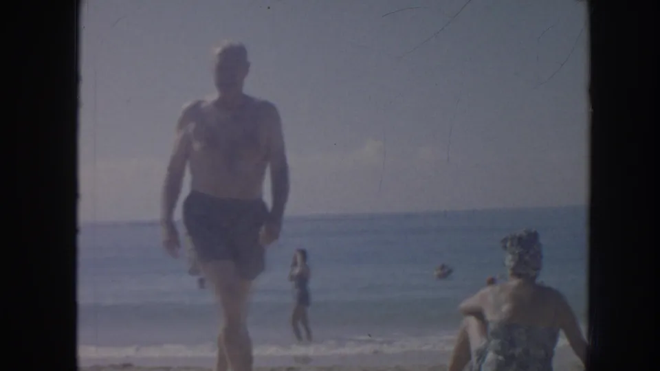 1961 Hawaii home-movie frame showing a man walking at the shoreline, with swimmers, surf, and beachgoers visible in soft 8mm focus.