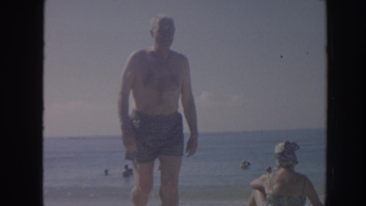 In this 1961 Hawaii 8mm frame, a shirtless man in patterned swim trunks approaches the camera near shallow surf and other beachgoers.