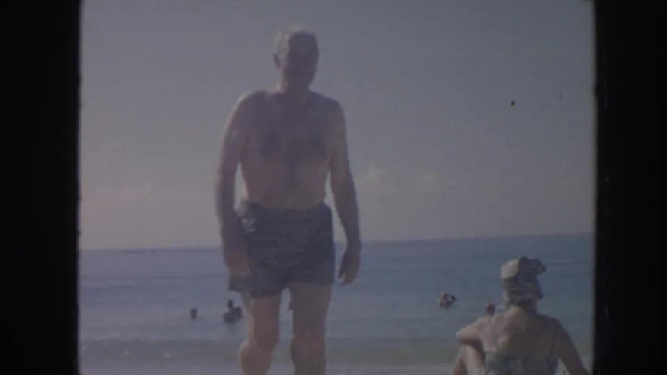 In this 1961 Hawaii 8mm frame, a shirtless man in patterned swim trunks approaches the camera near shallow surf and other beachgoers.