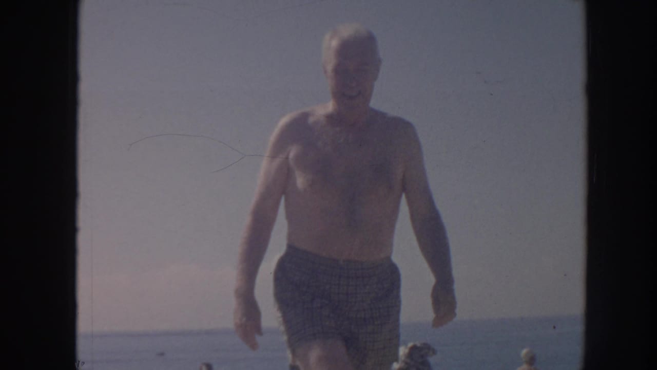 The man walks closer in the 1961 Hawaii beach footage, framed against bright sky, calm water, and distant beach activity.
