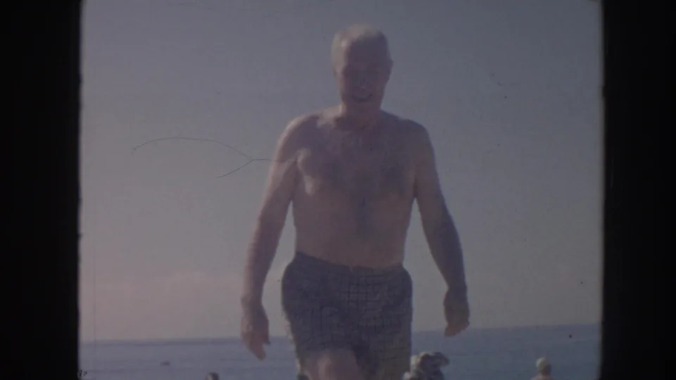 The man walks closer in the 1961 Hawaii beach footage, framed against bright sky, calm water, and distant beach activity.