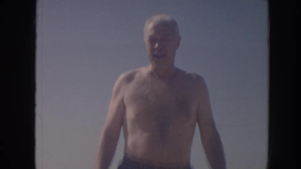 A closer 1961 Hawaii home-movie frame shows the man mid-stride in swim trunks, softly focused against sky and beach light.