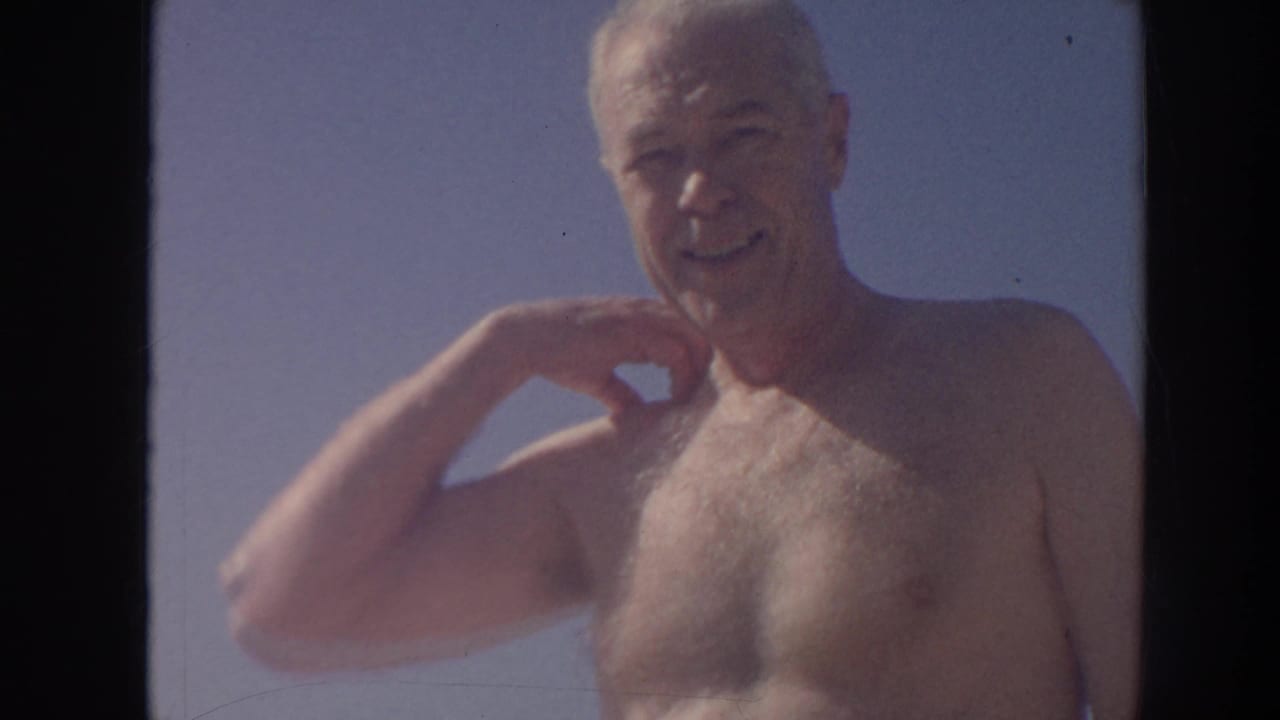 In 1961 Hawaii home-movie footage, an older shirtless gray-haired man smiles toward the camera in bright sun.