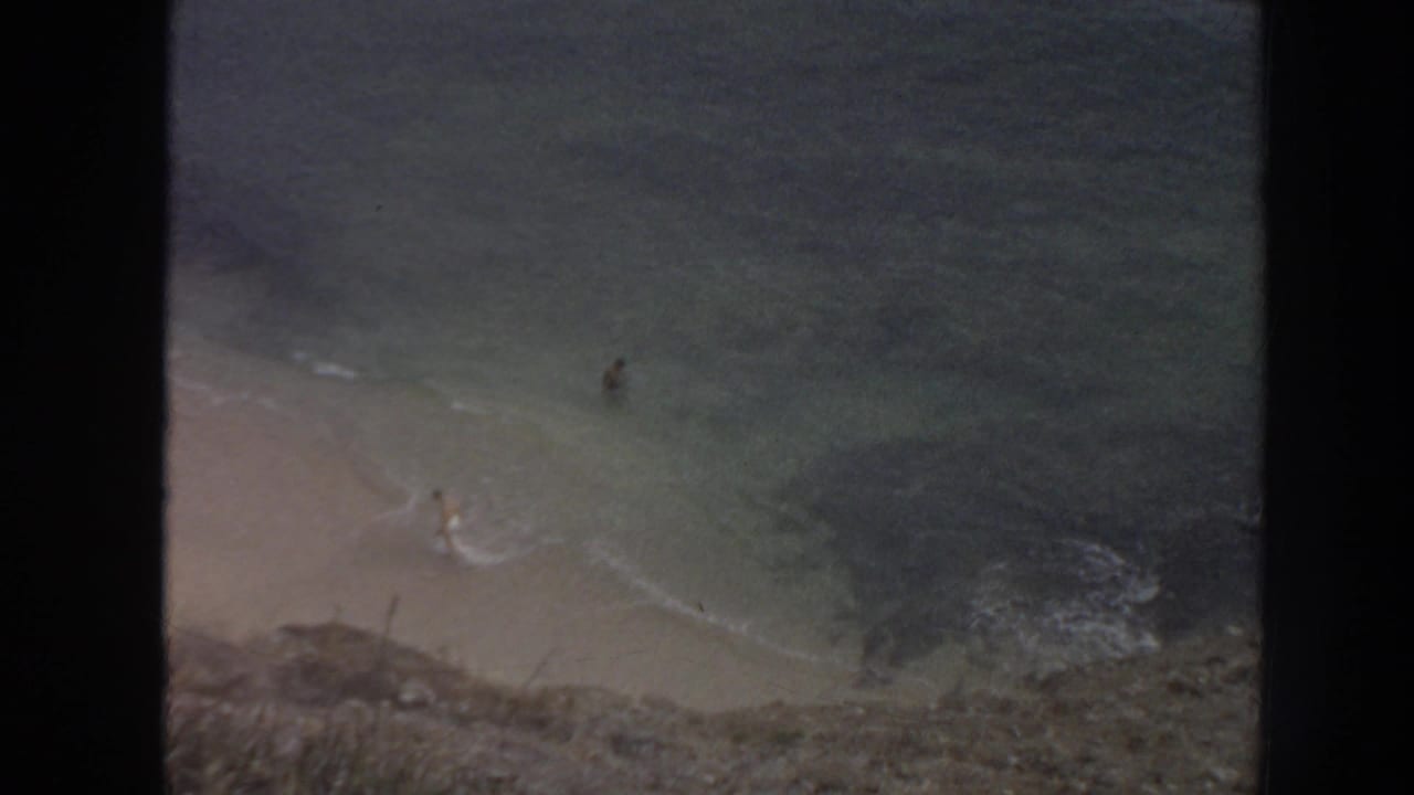 High view of a sandy beach and turquoise water below a rocky cliff with small swimmers visible.