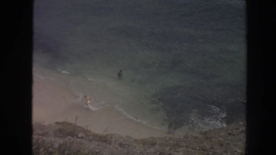 High-angle 1961 Hawaii beach view shows two small figures near the surf below a rocky cliff edge.