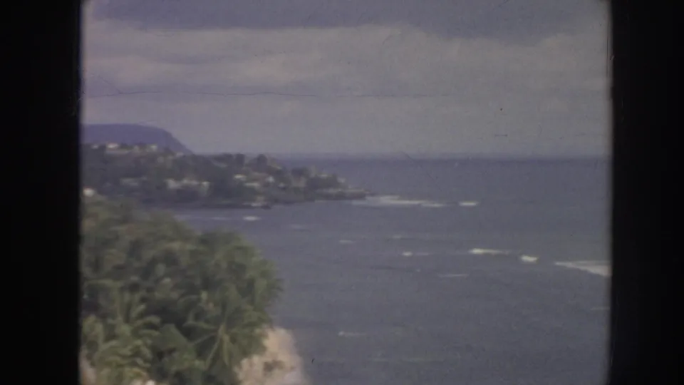 1961 Hawaii 8mm view of a palm-covered shoreline point, sandy beach edge, offshore surf, and distant hills across the water.