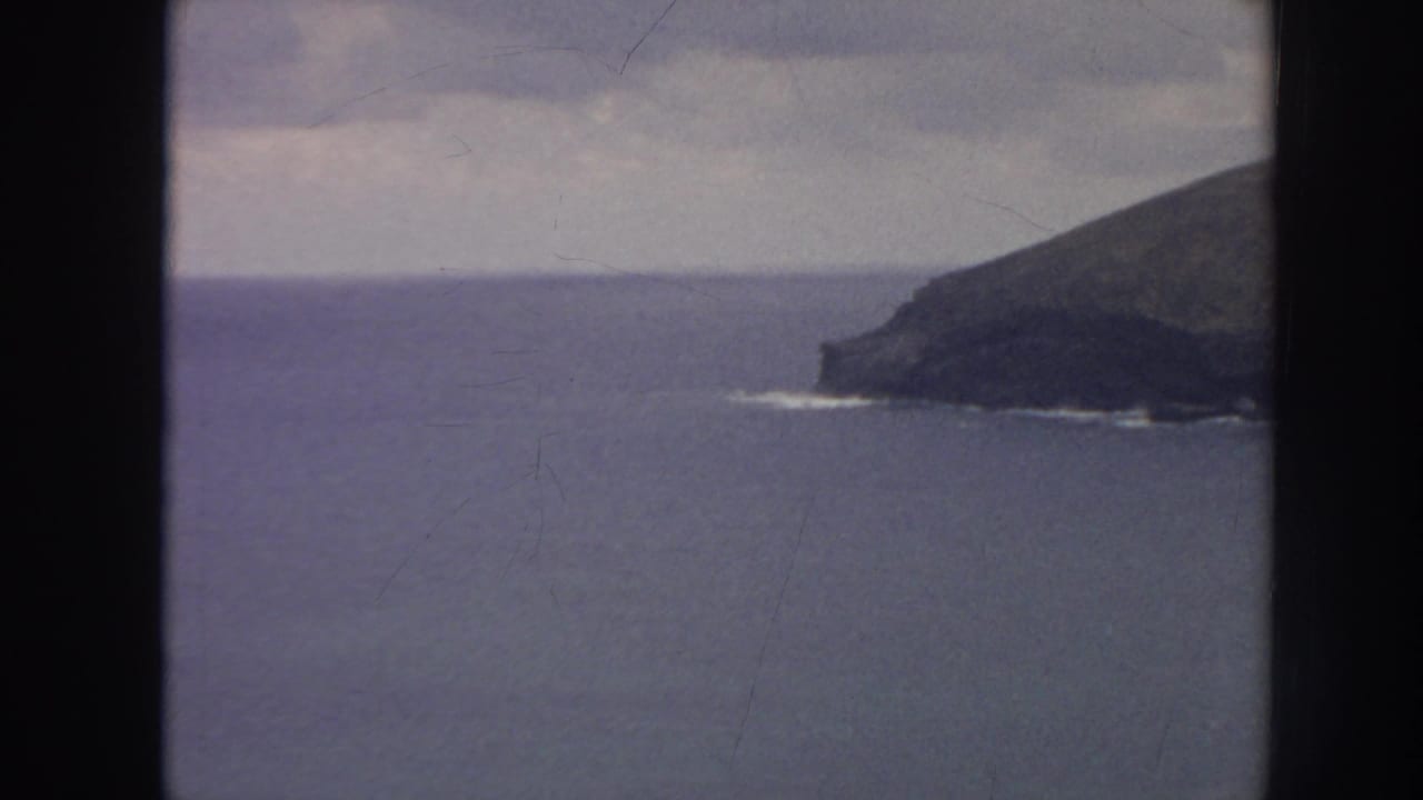 1961 Hawaii 8mm frame with open blue water, a dark rocky headland, faint surf at the base, and cloudy horizon.