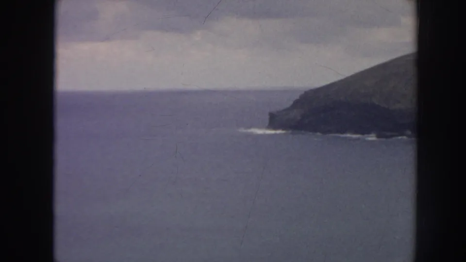 1961 Hawaii 8mm frame with open blue water, a dark rocky headland, faint surf at the base, and cloudy horizon.