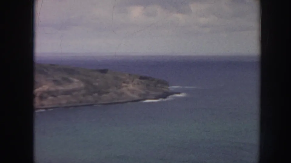 1961 Hawaii 8mm frame showing a rocky coastal point, blue sea, faint surf along the shore, and a hazy cloudy horizon.