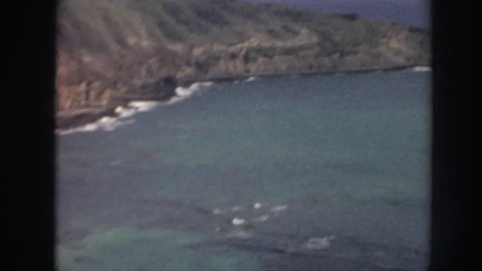 1961 Hawaii 8mm frame showing turquoise nearshore water, steep rocky coastline, and small white waves near the rocks.