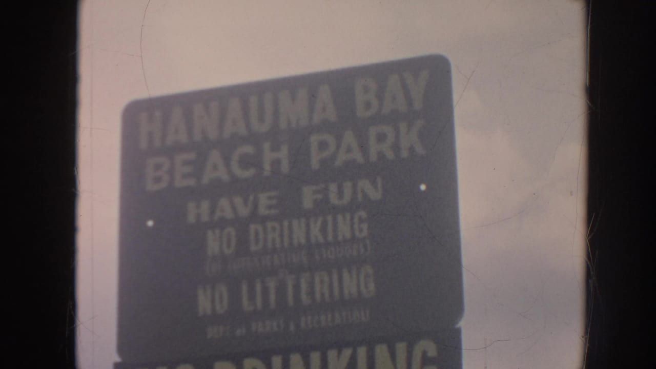 1961 Hawaii home-movie frame showing a dark Hanauma Bay Beach Park rules sign with cloudy sky behind it.
