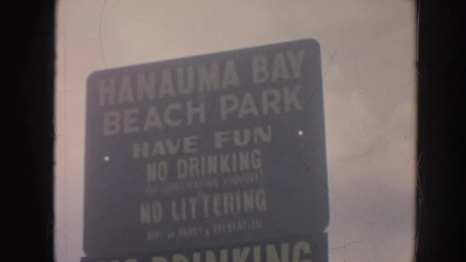 1961 Hawaii home-movie frame showing a dark Hanauma Bay Beach Park rules sign with cloudy sky behind it.
