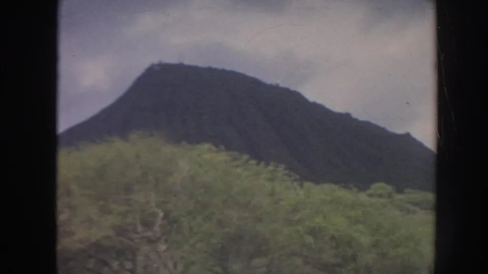 1961 Hawaii home-movie view of a dark mountain ridge rising above green vegetation under a cloudy sky.