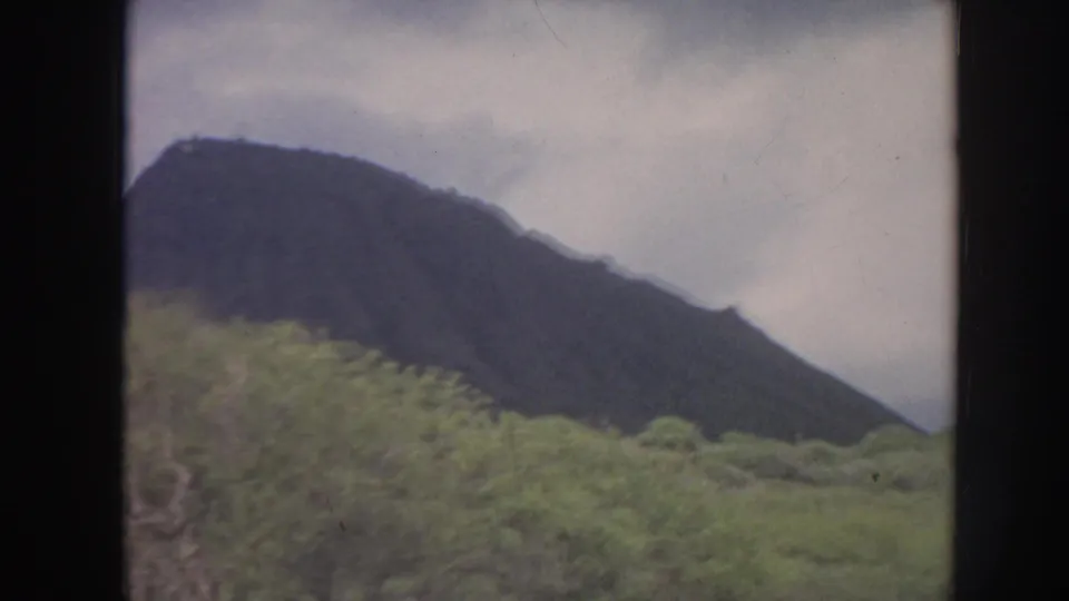 1961 Hawaii home-movie frame with a sloping mountain ridge, green brush, and cloudy sky.