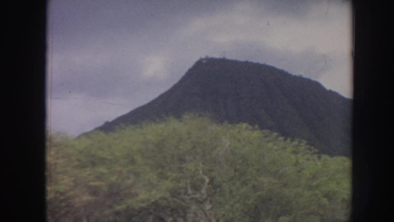 1961 Hawaii 8mm frame showing a dark mountain rising behind dense green vegetation beneath a cloudy sky.