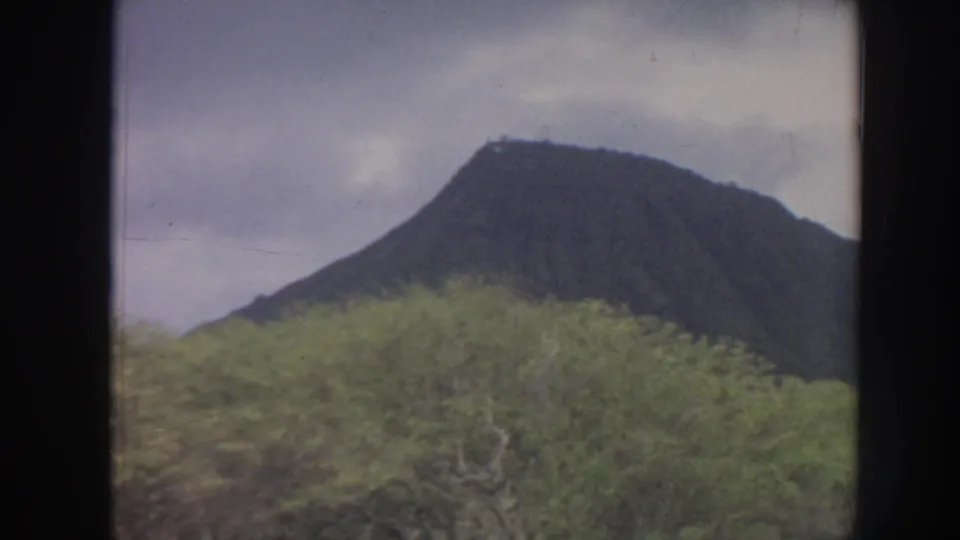 1961 Hawaii 8mm frame showing a dark mountain rising behind dense green vegetation beneath a cloudy sky.