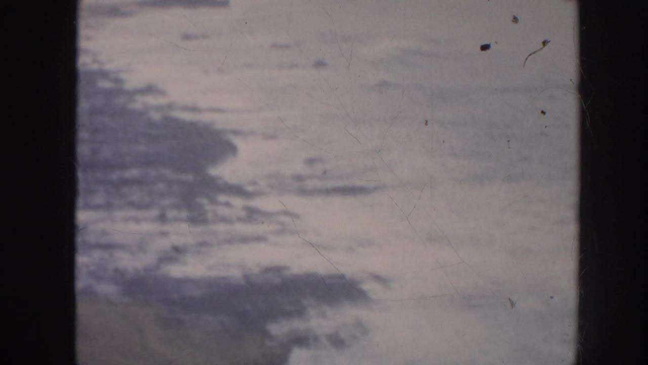 1961 Hawaii 8mm frame showing rough ocean water near a dark rocky shoreline under muted light.