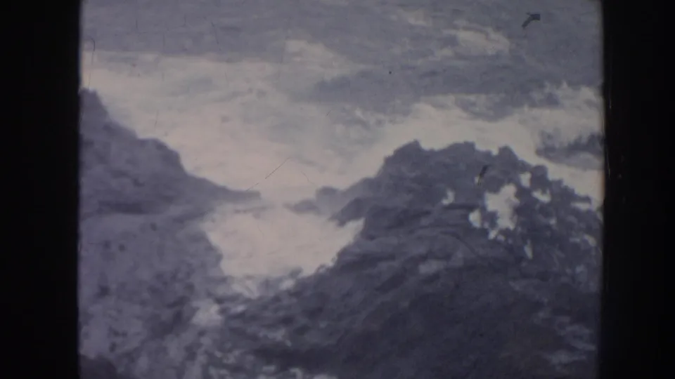 1961 Hawaii coastal frame showing white surf breaking among dark jagged rocks under a muted sky.