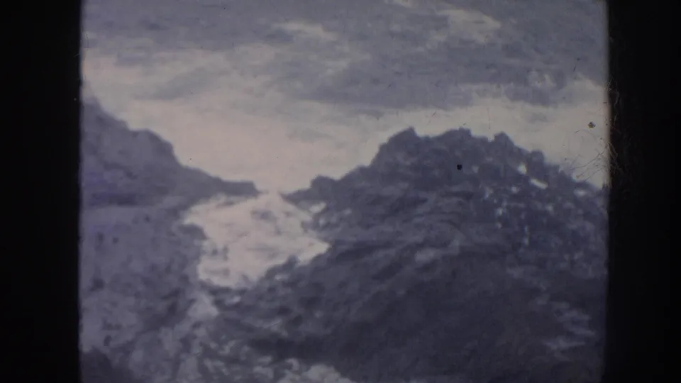 1961 Hawaii 8mm frame showing foamy waves striking jagged coastal rocks beneath an overcast sky.