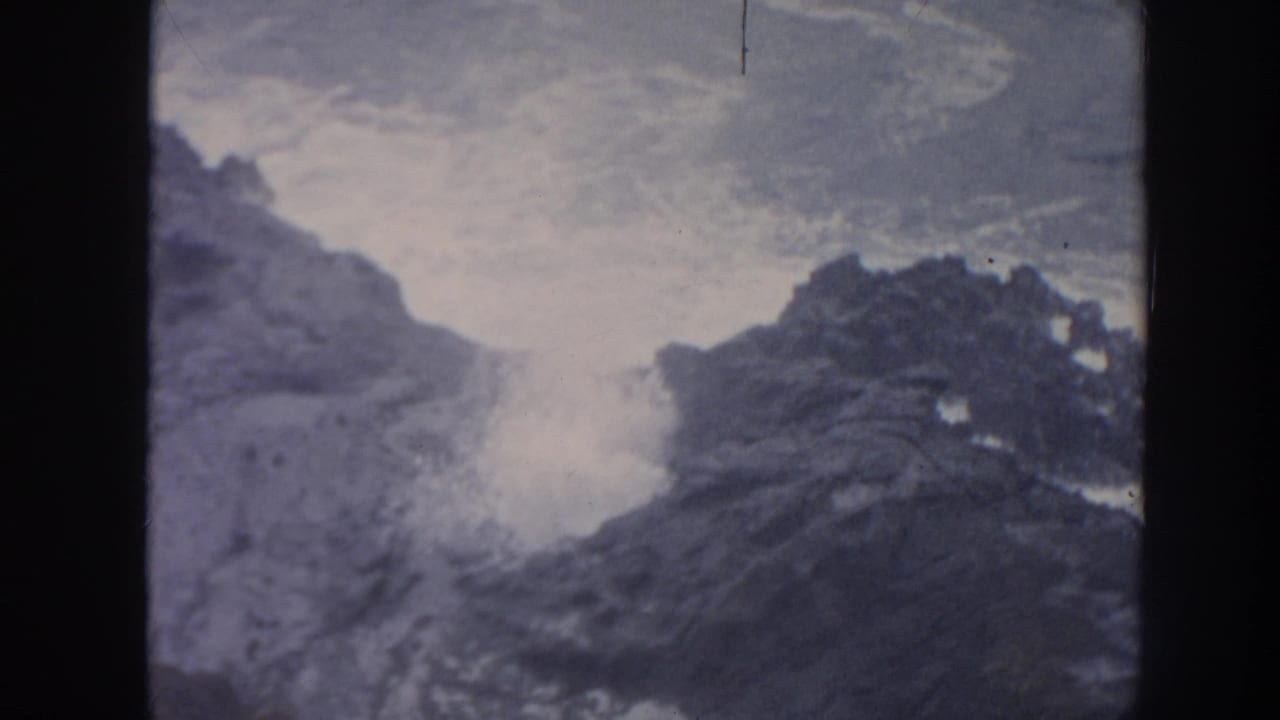A burst of white spray rises between dark rocks along the coast.