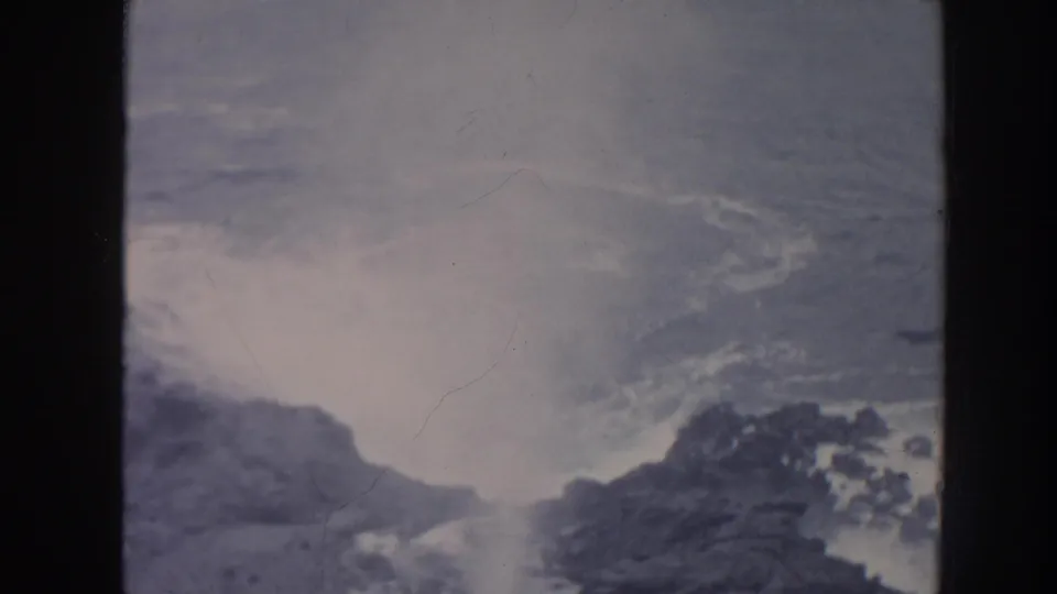 Vintage 8mm view of surf washing over Hawaii coastal rocks, with muted exposure and visible film texture.