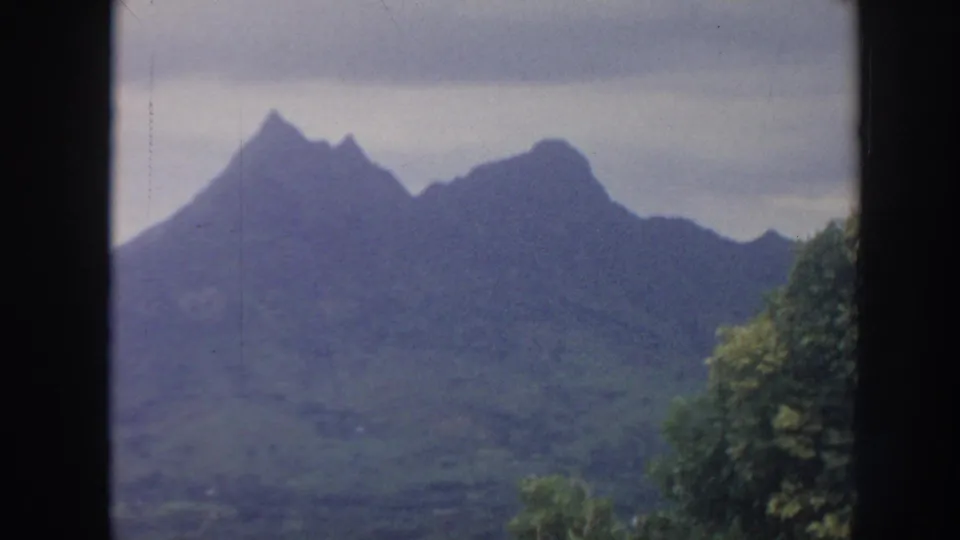 1961 Hawaii 8mm still showing a dark green mountain ridge, cloudy sky, and leafy foreground vegetation.