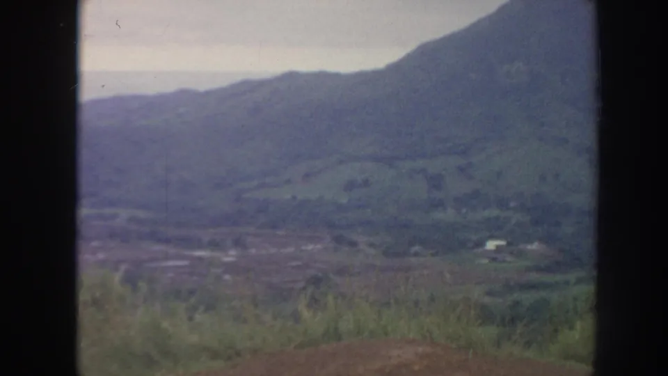1961 Hawaii home-movie frame of a broad green valley below mountain slopes, with foreground grasses and a small pale structure in the distance.