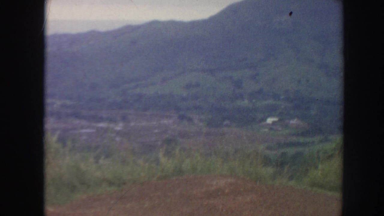 1961 Hawaii home-movie frame showing a hazy mountain valley from a high lookout, with foreground grass and dark film edges.