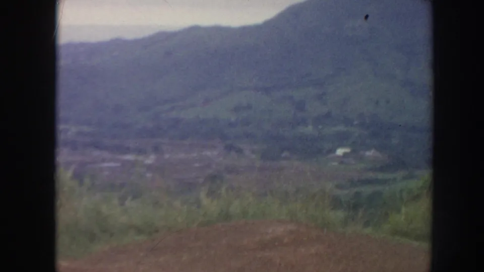 1961 Hawaii home-movie frame showing a hazy mountain valley from a high lookout, with foreground grass and dark film edges.