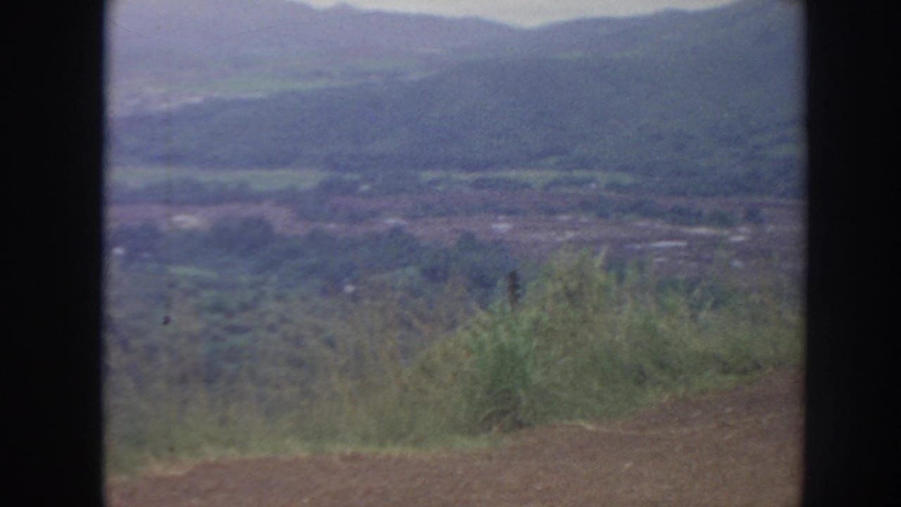 Mountain valley landscape with foreground vegetation and distant hills under a hazy sky.