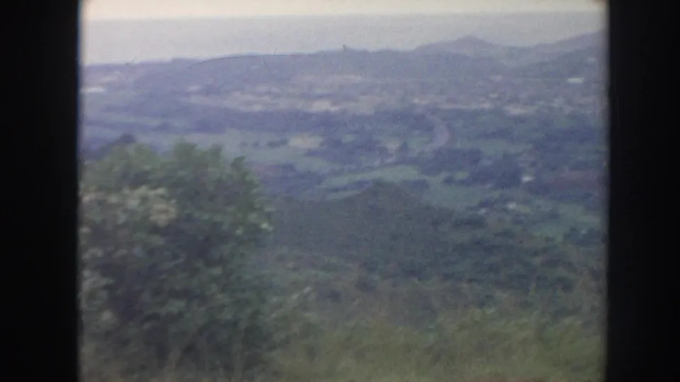 1961 Hawaii home-movie frame showing a wide green valley, foreground trees, and distant low mountain ridges.