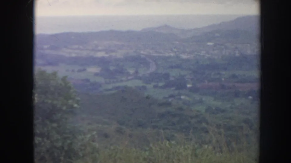 1961 Hawaii 8mm frame looking over a valley with a winding road, scattered pale structures, and hazy mountains.