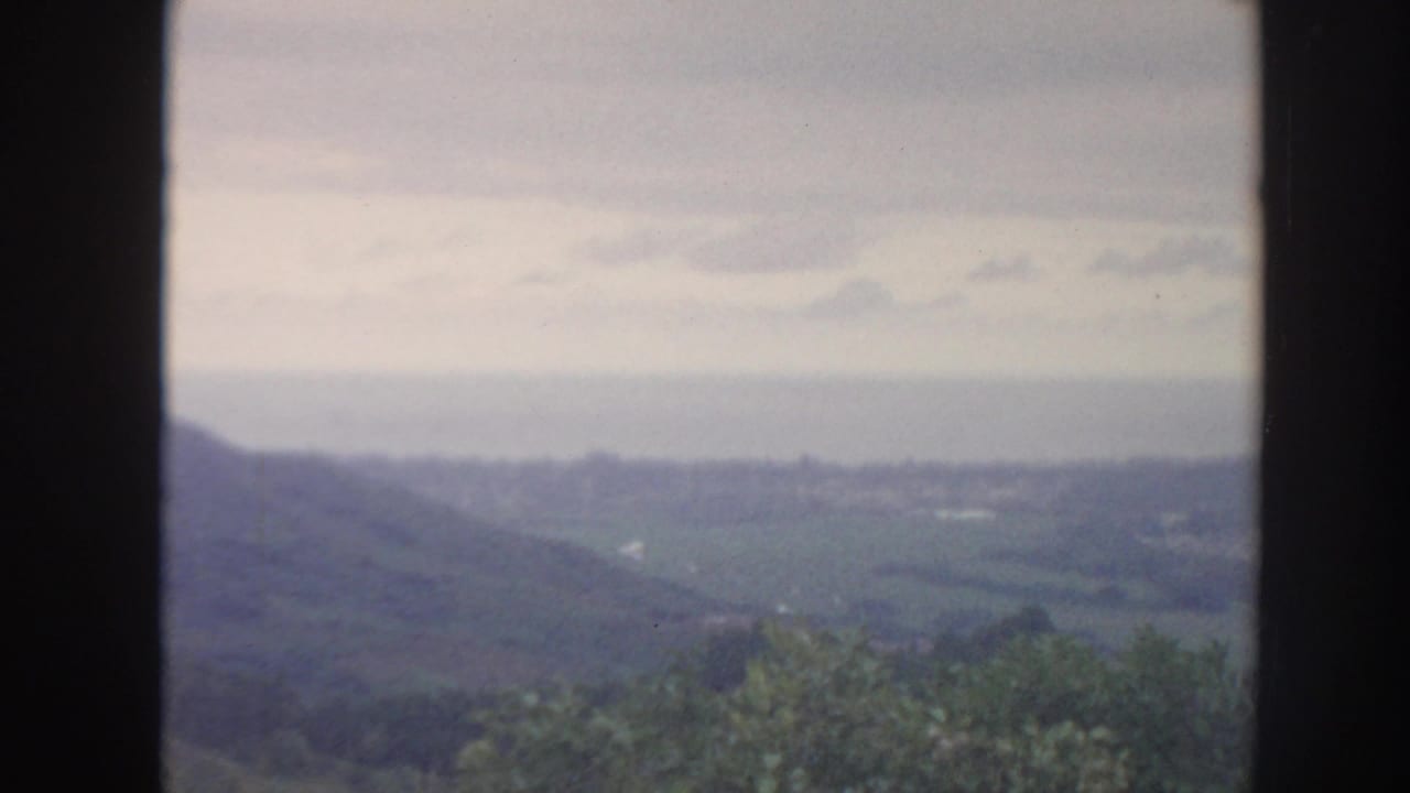 1961 Hawaii 8mm frame showing a hazy green valley, distant hills, and cloudy sky seen through a dark window frame.