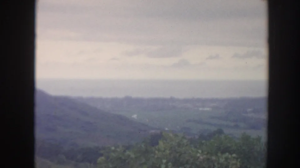 1961 Hawaii 8mm frame showing a hazy green valley, distant hills, and cloudy sky seen through a dark window frame.