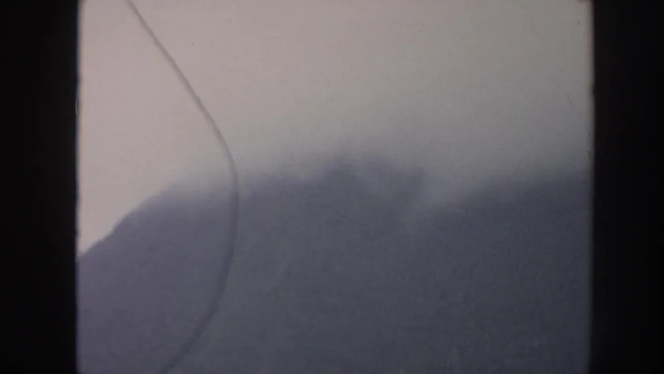 Muted 8mm frame of a rounded Hawaiian mountain silhouette below pale cloud cover, crossed by a thin scratch mark.