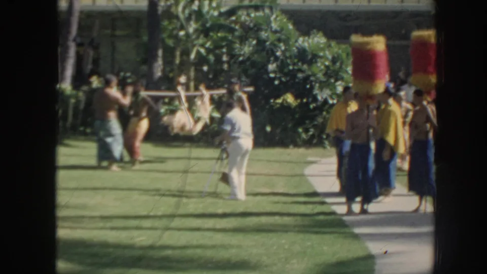 Adults at an outdoor Hawaii gathering carry a pig tied to a long pole across a lawn, with palms and greenery nearby.