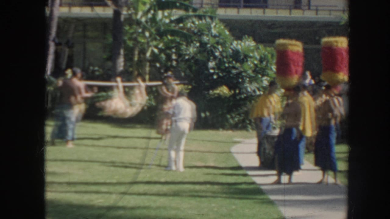 Performers in tall headpieces and yellow capes stand near a pig carried on a pole on a garden lawn.