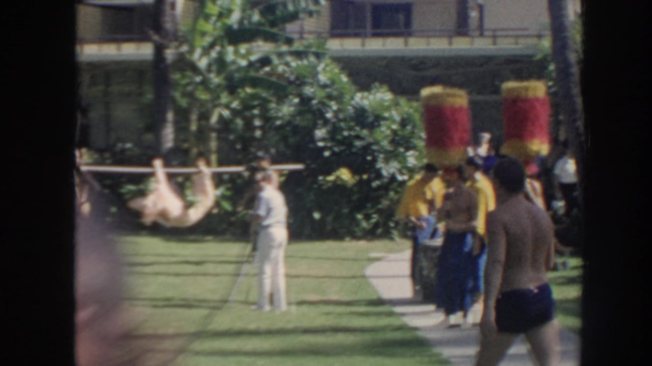 1961 Hawaii home-movie frame with a man in white clothing filming a garden performance as people in yellow capes and tall headpieces stand near a pig carried on a pole.
