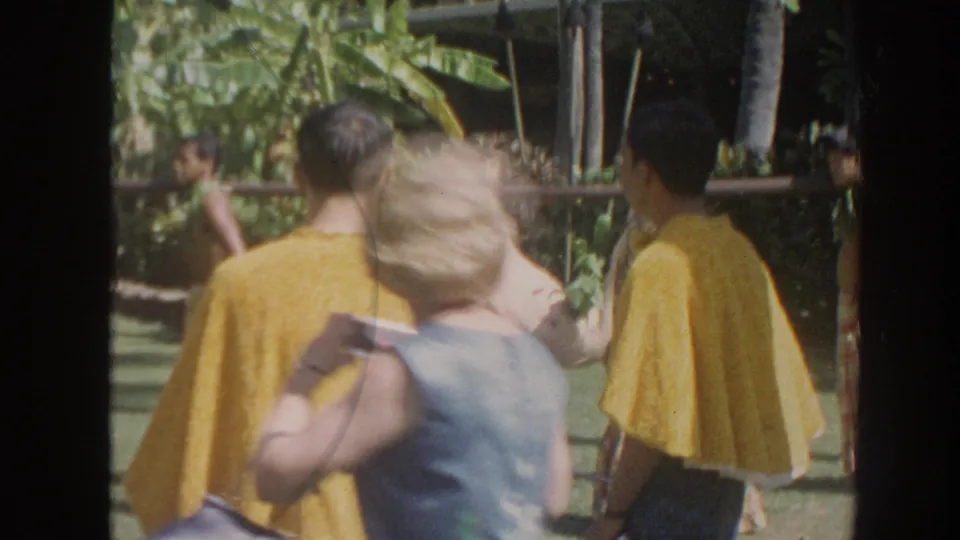 1961 Hawaii home-movie frame from behind spectators, showing performers in yellow capes near a pig suspended from a pole in a sunny tropical garden.