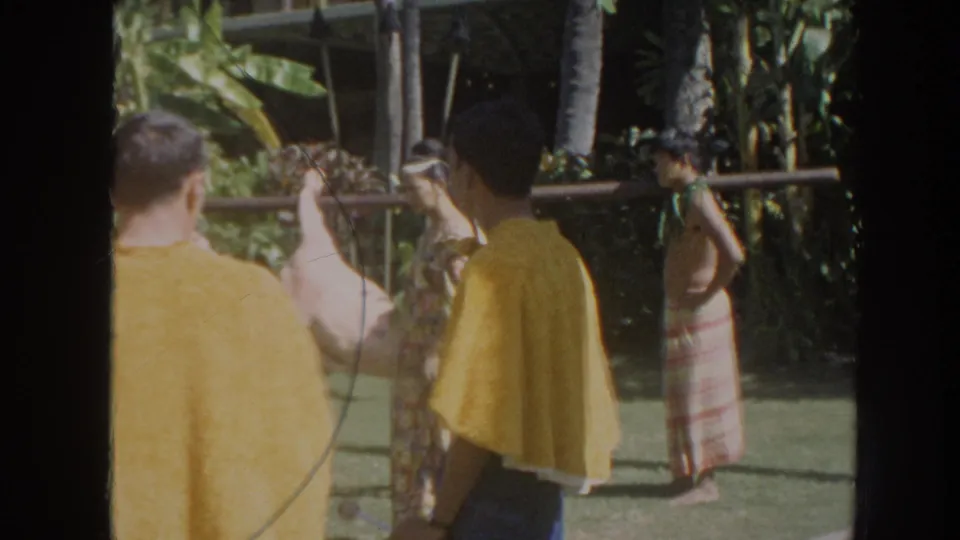 1961 Hawaii home-movie frame showing spectators and performers in yellow capes beside a pig carried on a pole, with a woman in a patterned skirt near the garden edge.