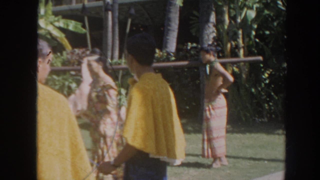 1961 Hawaii 8mm frame showing adults in patterned wraps gathered near a long pole in a lush outdoor garden.