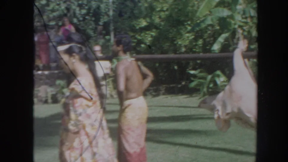 1961 Hawaii 8mm frame showing two adults in wrap garments near a suspended pig and dense tropical foliage.