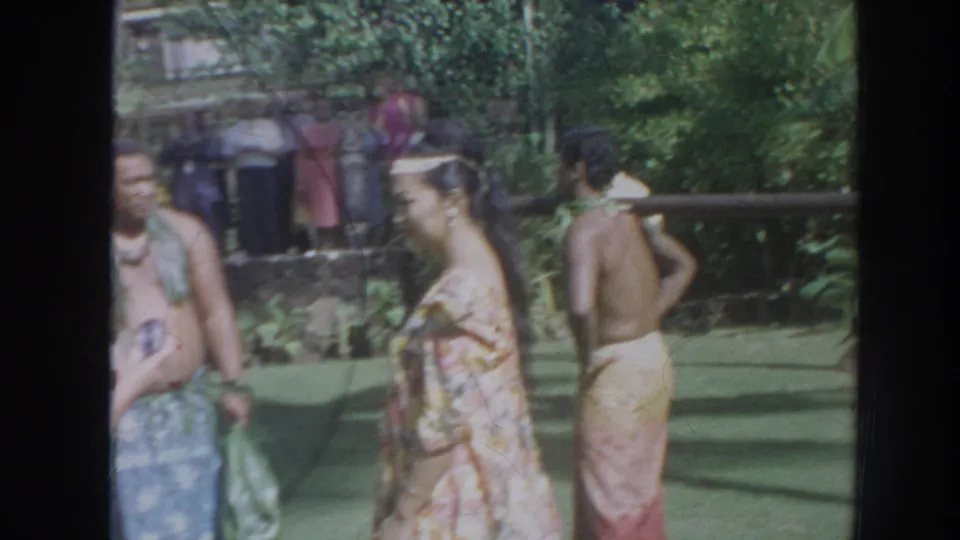 1961 Hawaii home-movie frame of adults in colorful wraps gathered near the pig roast setup in a leafy garden.