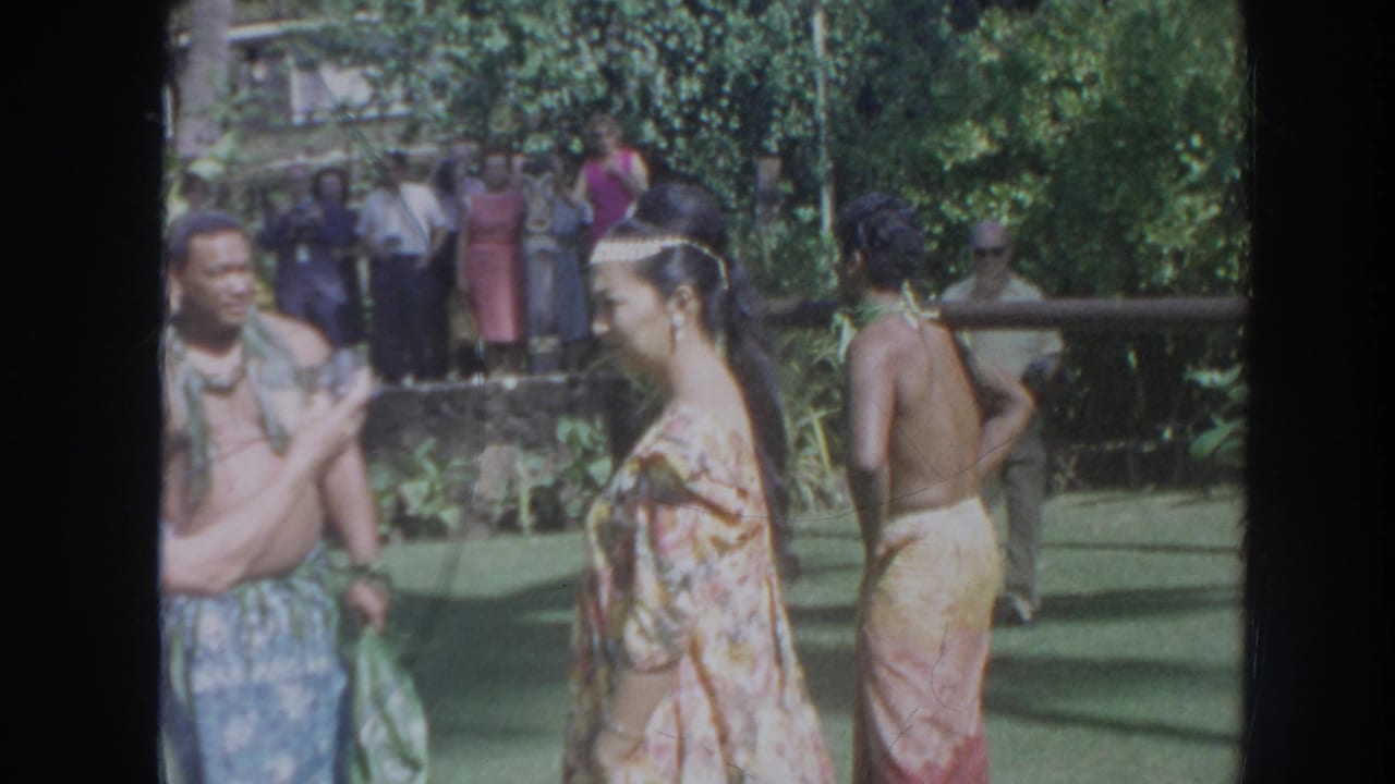 1961 Hawaii 8mm frame showing adults in ceremonial dress near a pole with a suspended pig, with onlookers in the background.