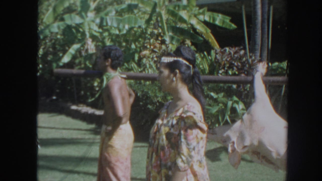 1961 Hawaii home-movie frame with two adults in tapa-style clothing near tropical foliage and a whole pig suspended from a pole.