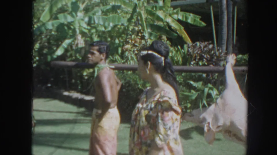 1961 Hawaii 8mm frame showing adults in ceremonial clothing beside a rail, with a whole pig hanging from a pole at the edge of the shot.