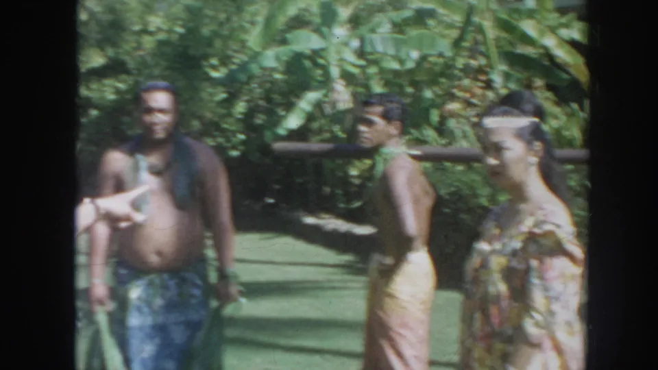 1961 Hawaii home-movie frame with adults in ceremonial dress moving through a tropical garden as a man at left faces the camera.