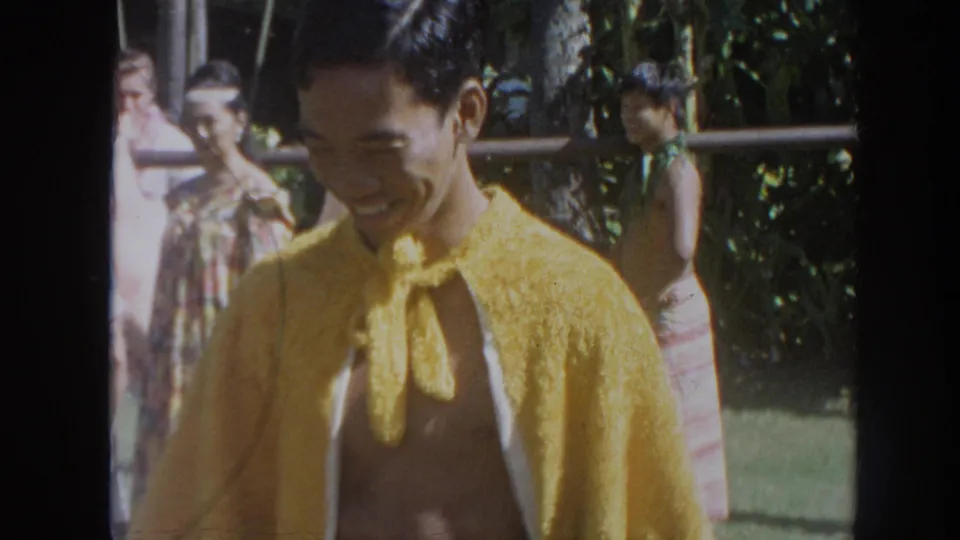 1961 Hawaii frame with a smiling man in a yellow woven-style cloak standing near other people in a luau setting.